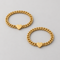 Two hands holding each other wearing gold bracelets with heart charms on a warm-toned background.