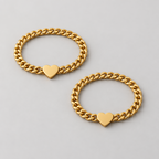 Two hands holding each other wearing gold bracelets with heart charms on a warm-toned background.